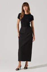 A woman is wearing a short-sleeved black lace top with intricate floral patterns, paired with a high-waisted, ankle-length black skirt that features a front button and hidden zipper. She completes her outfit with black open-toe high-heeled sandals.